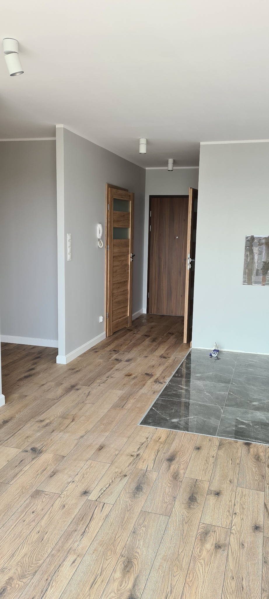 Wnętrze mieszkania po remoncie z drewnianą podłogą przechodzącą w ciemne płytki, widoczne dwie drewniane ościeżnice i szare ściany.