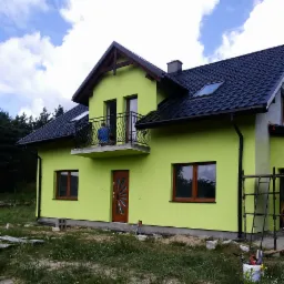 Nowo otynkowany dom jednorodzinny w odcieniu limonki z ciemną dachówką, balkonem z ozdobną balustradą i drewnianą stolarką okienną; na balkonie widoczna osoba w niebieskiej koszulce, przy ścianie...