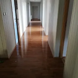 Długi korytarz z drewnianą podłogą i boazerią na ścianach, widok w perspektywie z otwartymi drzwiami po lewej stronie i szeregiem drzwi po obu stronach, oświetlony wiszącymi lampami.