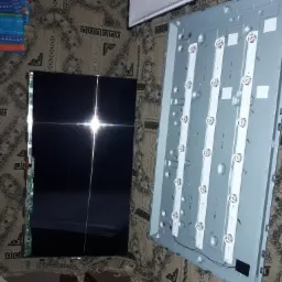 Rozłożony telewizor: matryca z odbiciem światła i panel tylny z listwami LED, leżące na wzorzystej tkaninie.