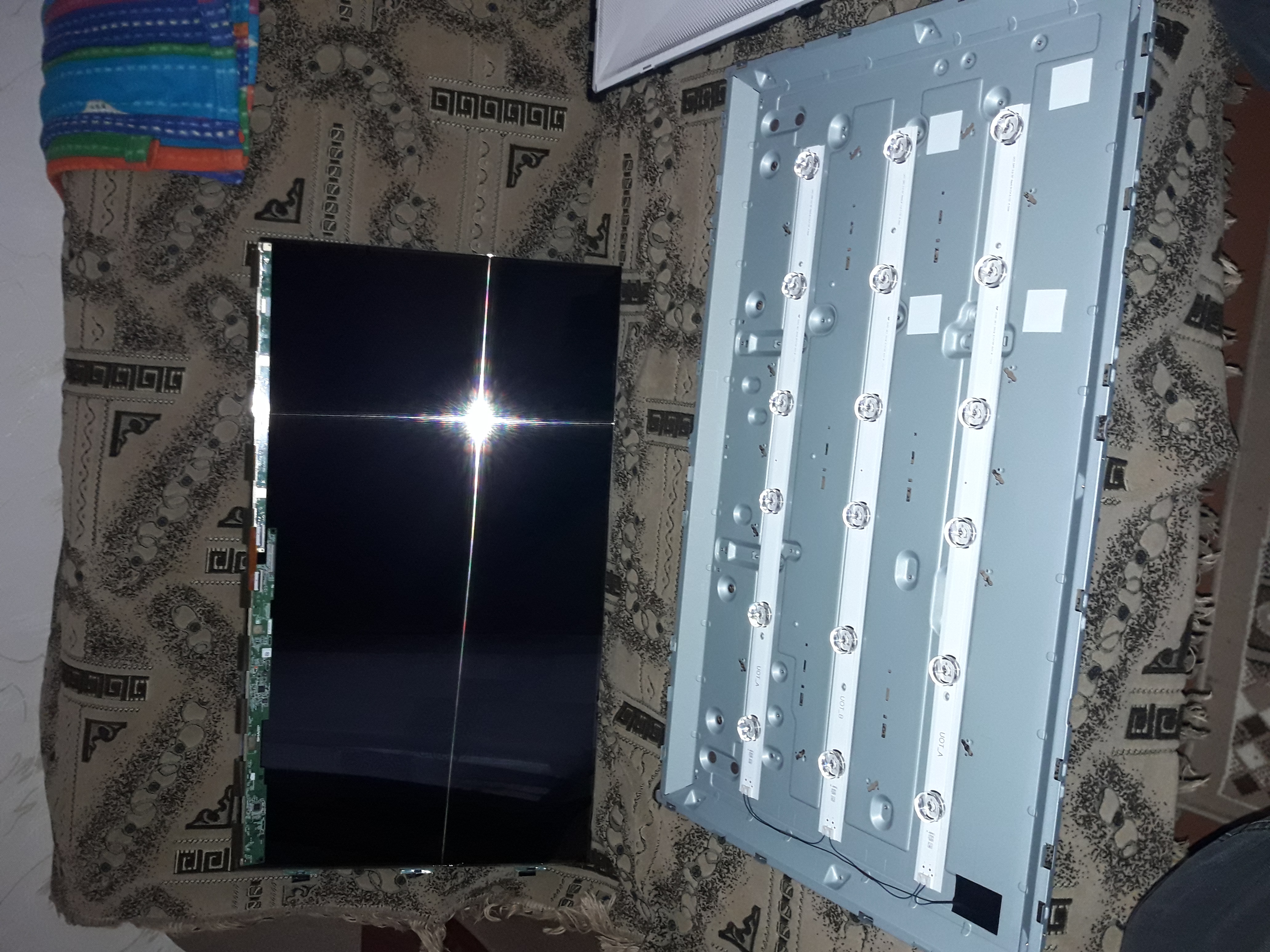 Rozłożony telewizor: matryca z odbiciem światła i panel tylny z listwami LED, leżące na wzorzystej tkaninie.