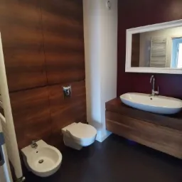 Nowoczesna łazienka z drewnianą zabudową, wiszącą toaletą i bidetem, umywalką nablatową oraz dużym lustrem w białej ramie na bordowej ścianie.