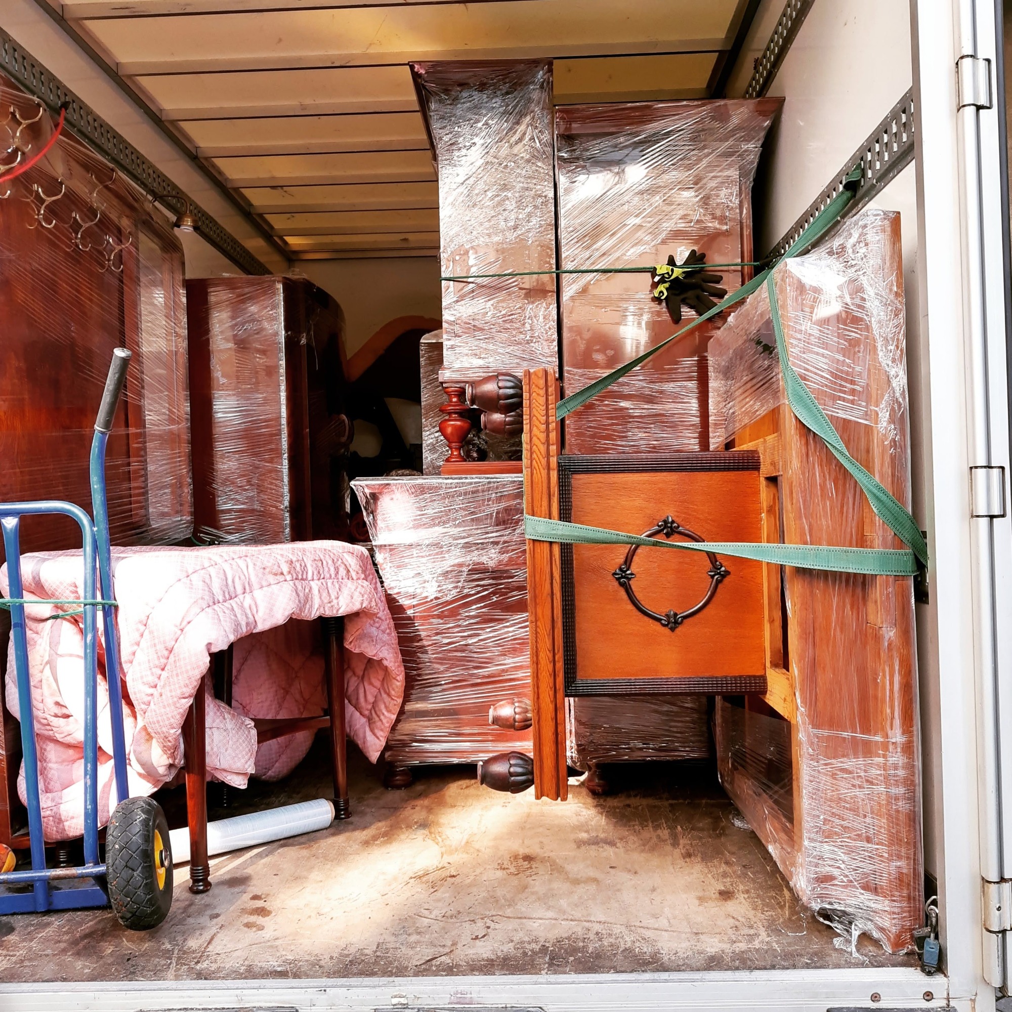Wnętrze furgonetki wypełnionej antycznymi meblami zabezpieczonymi folią stretch i pasami transportowymi, widoczny wózek transportowy z różową kołdrą.
