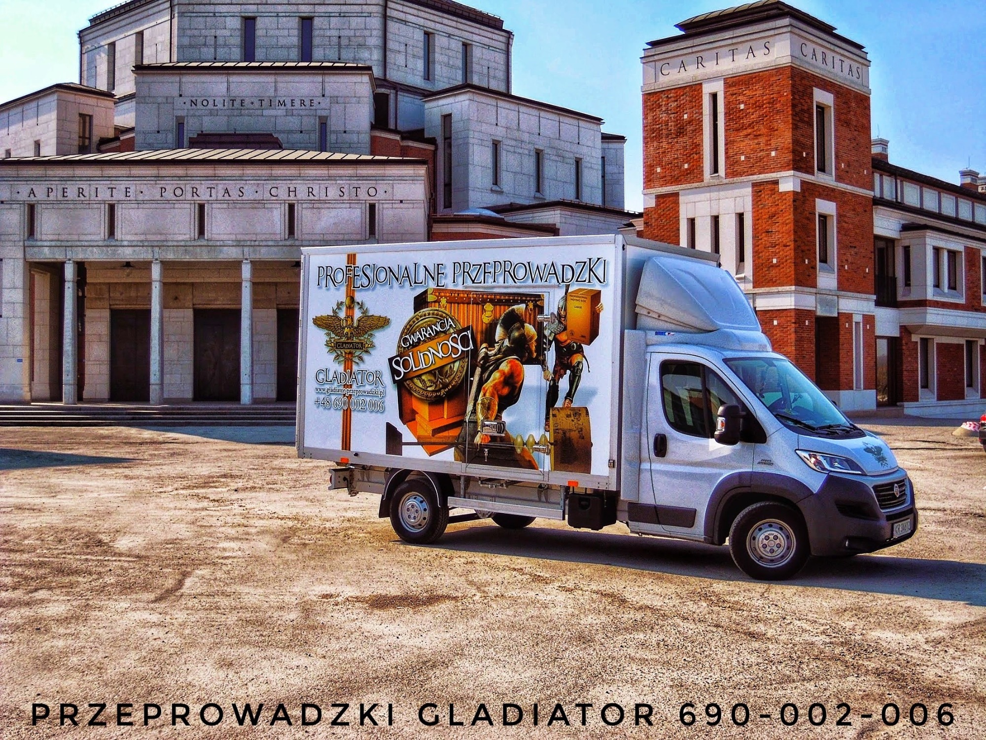 Biała furgonetka z reklamą firmy oferującej usługi transportowe, na której widnieje wizerunek gladiatora i hasło 'Profesjonalne Przeprowadzki', zaparkowana na placu przed budynkiem o architekturze...