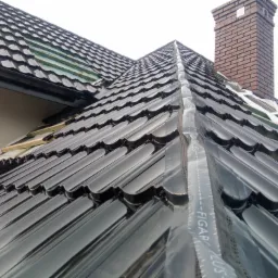 Montaż błyszczącej, czarnej dachówki ceramicznej na dachu domu jednorodzinnego, widoczny komin z cegły i folia ochronna na połaci.