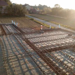 Zbrojenie płyty fundamentowej z widocznymi butelkami z wodą używanymi do poziomowania, w tle krajobraz z drogą i domami w promieniach wschodzącego słońca.