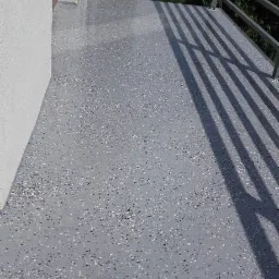 Szara posadzka z drobnymi, ciemnymi i jasnymi elementami na balkonie, widok na metalową barierkę i zieleń w tle.