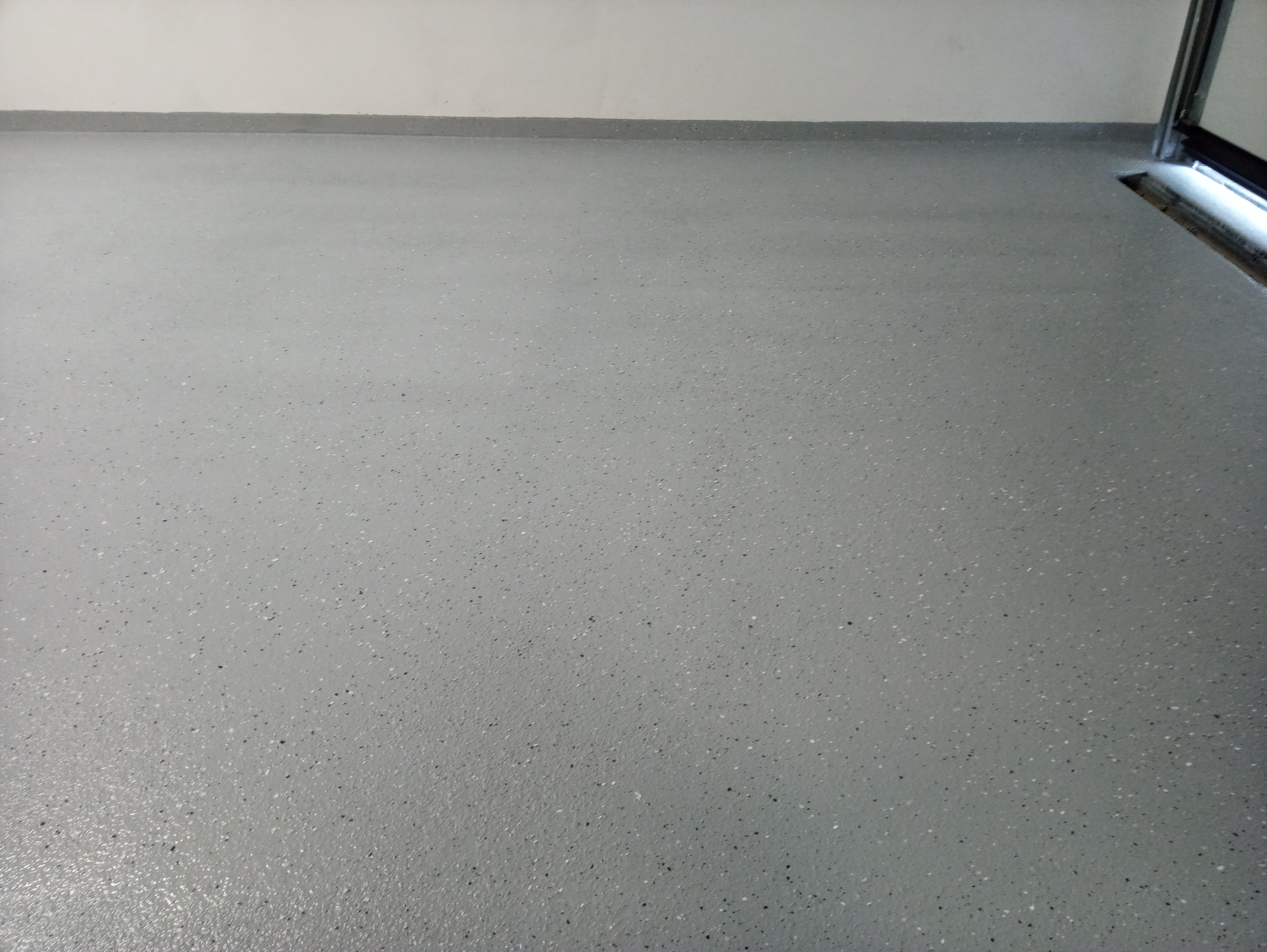 Jasnoszara, gładka podłoga z drobnymi, ciemnymi i jasnymi plamkami, widoczny fragment ściany i brama garażowa.