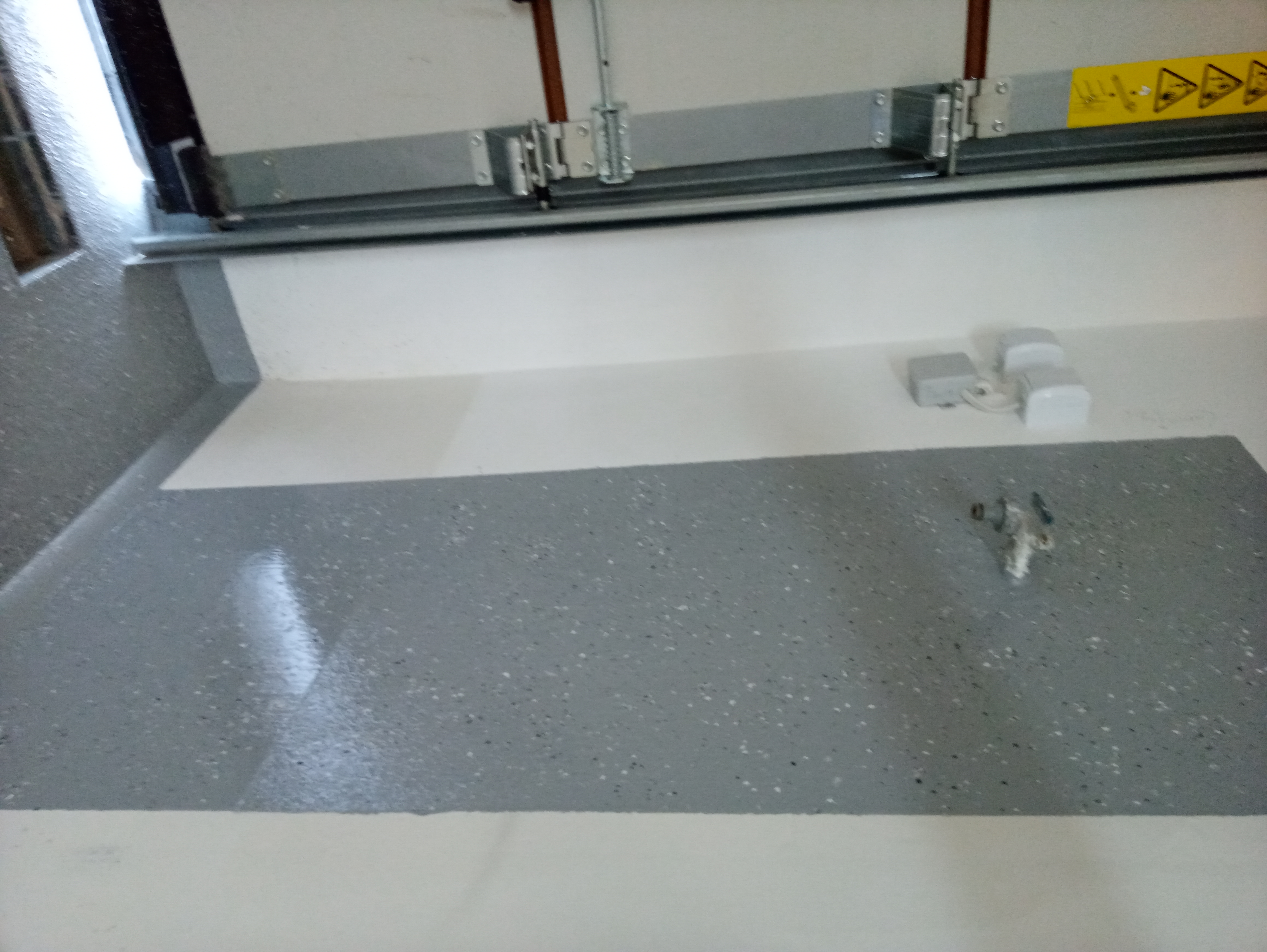 Szara, nakrapiana powierzchnia w garażu, kontrastująca z białą ścianą i fragmentem bramy garażowej z żółtą naklejką ostrzegawczą.