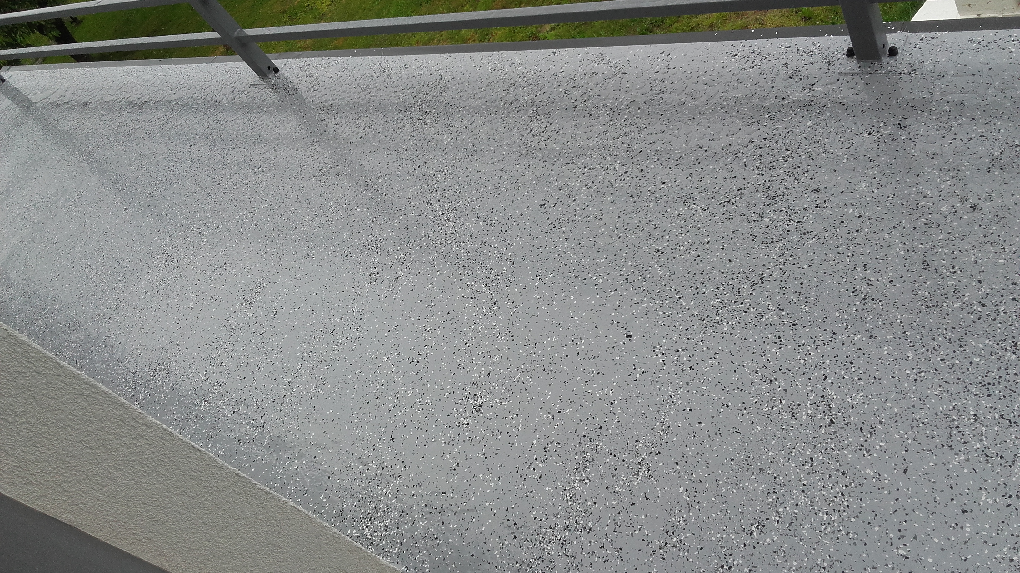 Szara, wykończona posadzka balkonowa z drobnymi, czarnymi i białymi wtrąceniami, widok z góry, fragment metalowej barierki w tle.