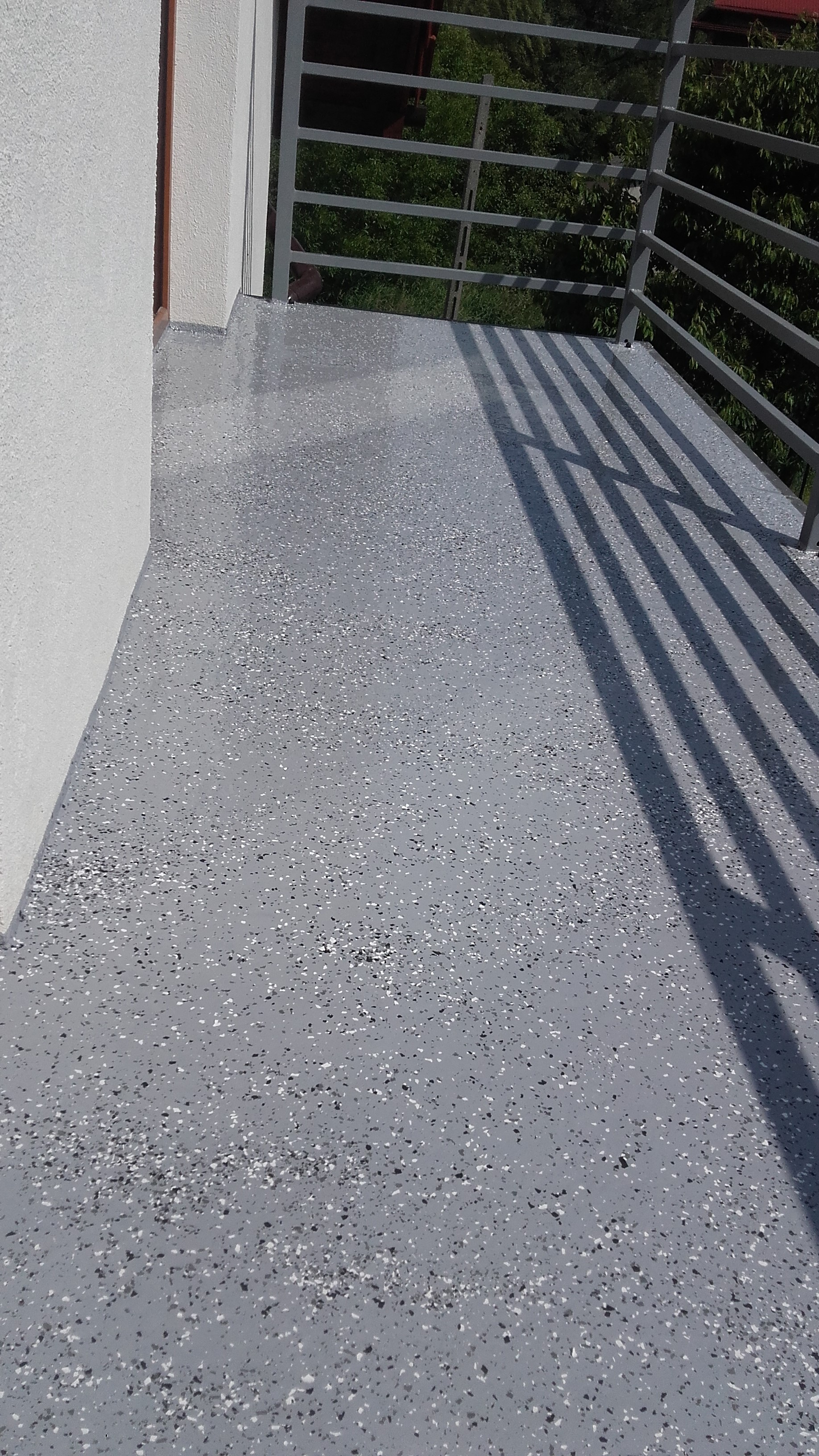 Szara posadzka z drobnymi, ciemnymi i jasnymi elementami na balkonie, widok na metalową barierkę i zieleń w tle.