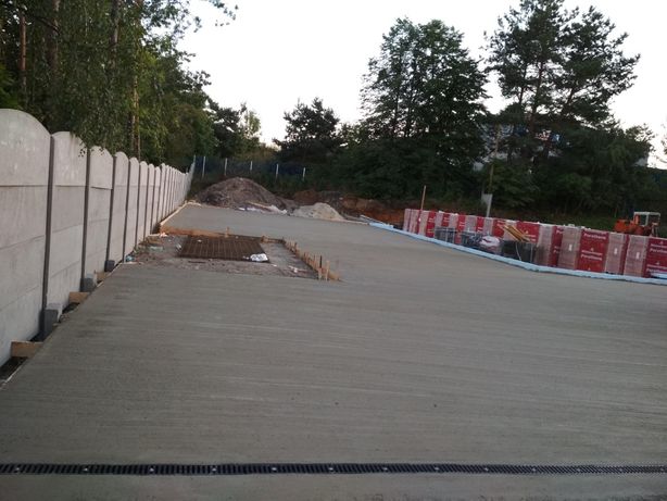 Wylewka betonowa na przygotowanym terenie budowy, widoczna siatka zbrojeniowa, szalunki, palety z materiałami budowlanymi oraz betonowy płot w tle.