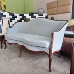 Jasnoszara sofa w stylu retro z drewnianymi nogami, obok krzesło z jasnym obiciem, w tle ściana z panelami tapicerowanymi w różne wzory.