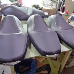 Cztery obszyte ciemnoszare siedzenia motocyklowe z czerwoną nicią, z metalowymi elementami z napisem Yamaha, leżące na stole w warsztacie, w tle maszyna do szycia i materiały.