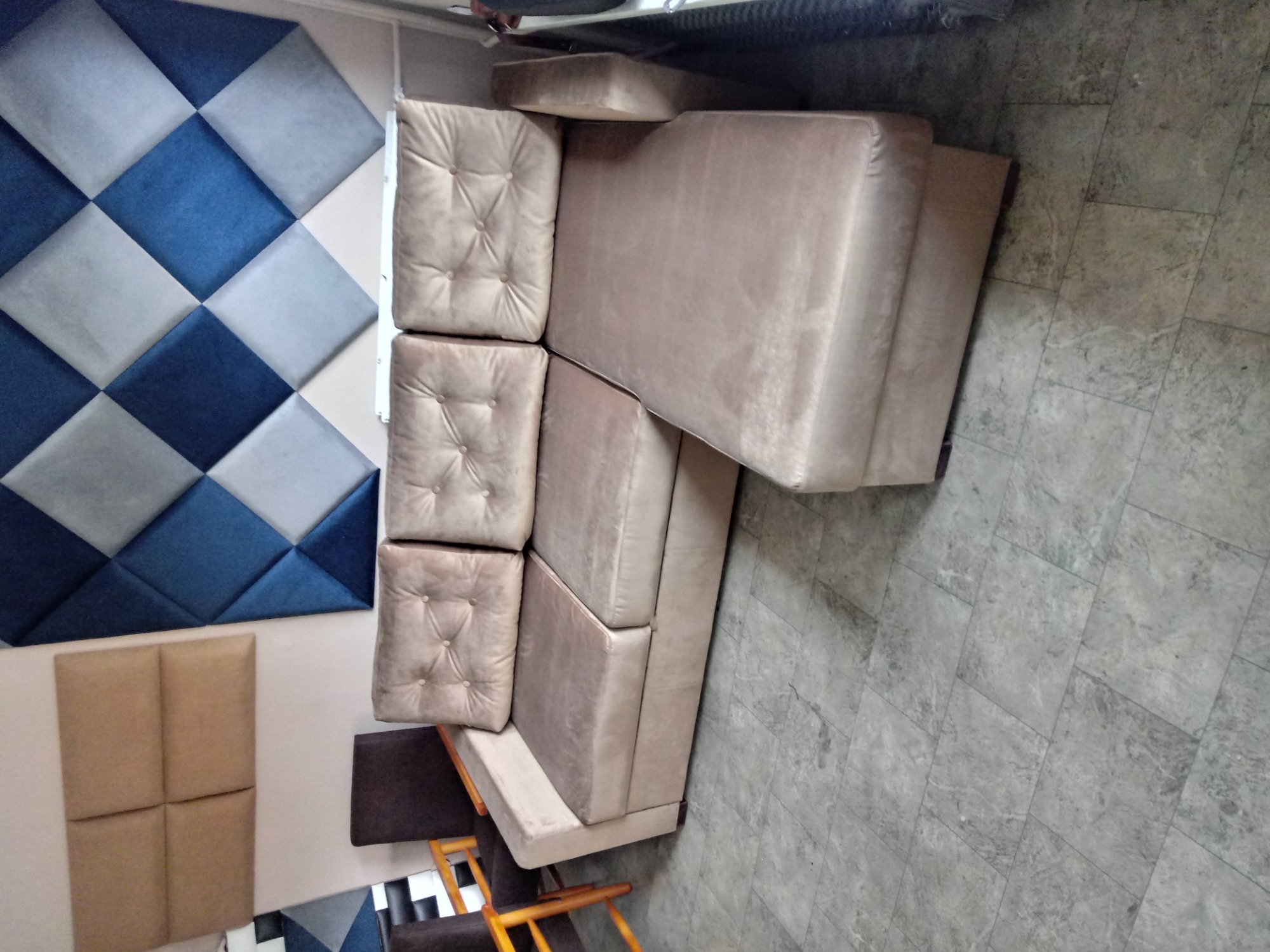 Beżowa sofa narożna z pikowanymi poduchami, ustawiona w pomieszczeniu z panelami tapicerowanymi na ścianie w odcieniach niebieskiego i beżu.