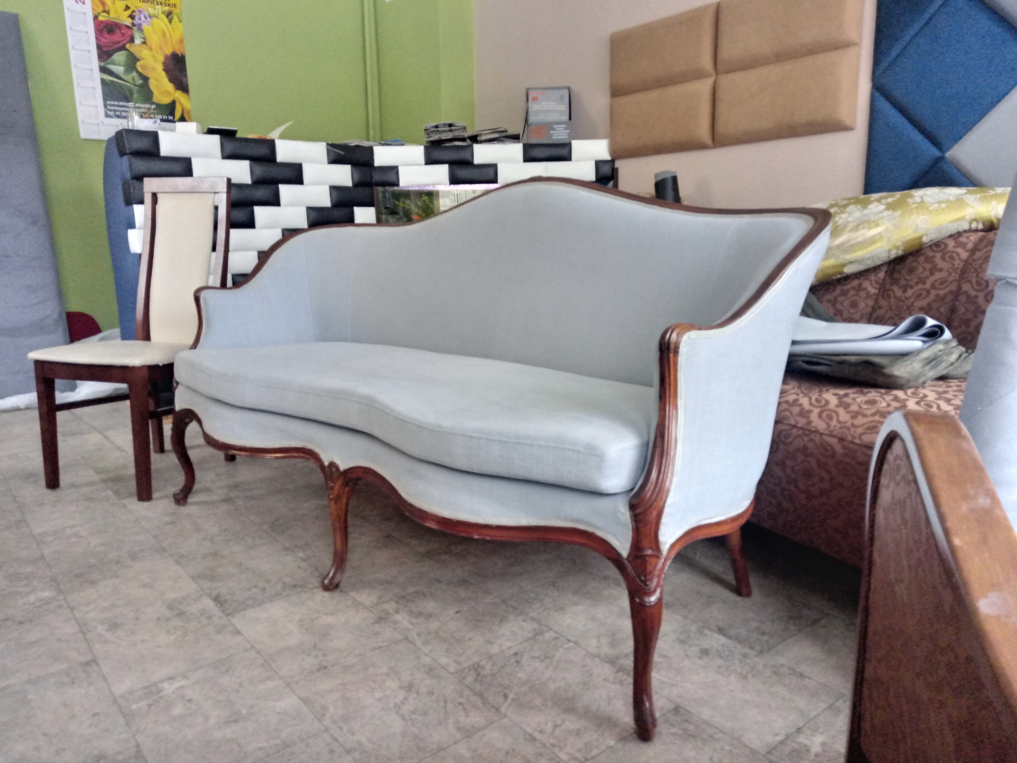 Jasnoszara sofa w stylu retro z drewnianymi nogami, obok krzesło z jasnym obiciem, w tle ściana z panelami tapicerowanymi w różne wzory.