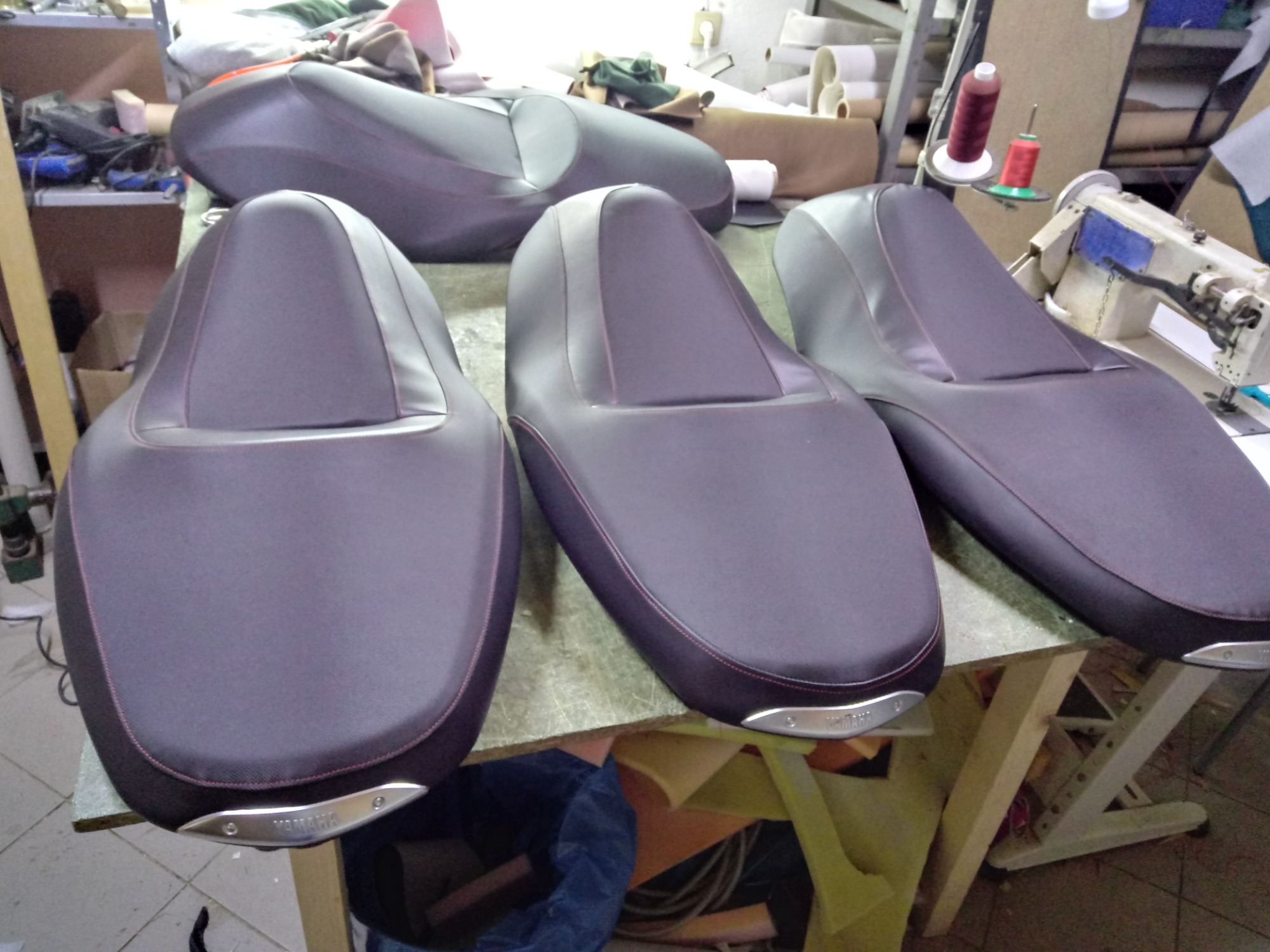 Cztery obszyte ciemnoszare siedzenia motocyklowe z czerwoną nicią, z metalowymi elementami z napisem Yamaha, leżące na stole w warsztacie, w tle maszyna do szycia i materiały.