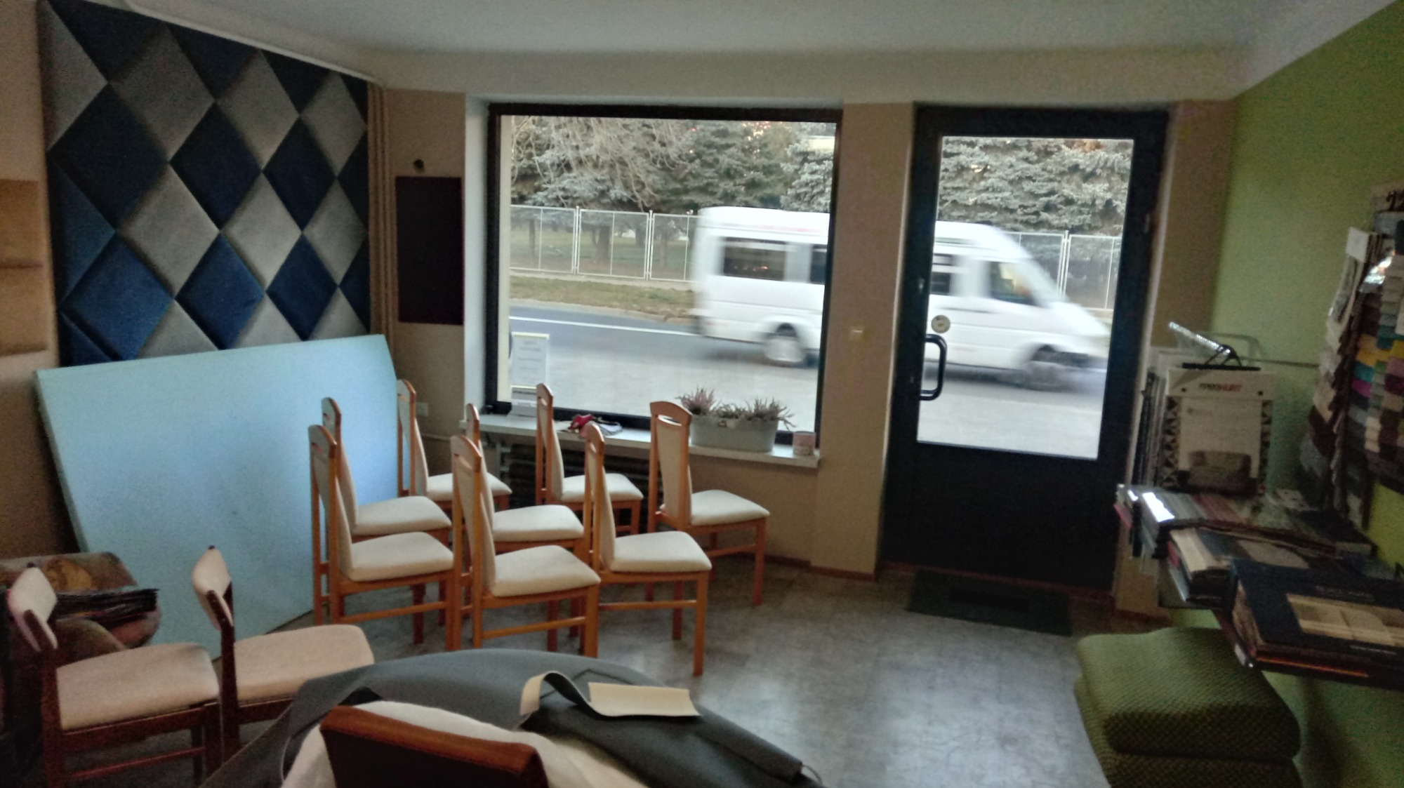 Wnętrze warsztatu z przygotowanymi krzesłami do obicia, w tle okno z widokiem na ulicę i przejeżdżający biały bus oraz panel tapicerowany na ścianie.