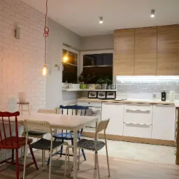 Wykończona kuchnia z białymi cegłami na ścianie, drewnianymi szafkami i białymi frontami, oświetleniem LED pod szafkami oraz stołem z krzesłami w różnych kolorach.