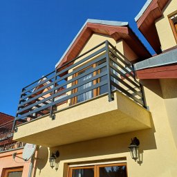 Andrzej Mazelon Usługi Spawalnicze - Balkon z nowoczesną, szarą balustradą z poziomych profili na tle błękitnego nieba i elewacji budynku w ciepłym odcieniu. Widoczne lampy zewnętrzne i fragment dachu.