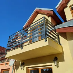 Balkon z nowoczesną, szarą balustradą z poziomych profili na tle błękitnego nieba i elewacji budynku w ciepłym odcieniu. Widoczne lampy zewnętrzne i fragment dachu.