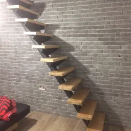 Schody wspornikowe z drewnianymi stopniami i metalowymi wspornikami, zamontowane przy szarej ścianie z cegły, widoczne gniazdko elektryczne i fragment sofy.