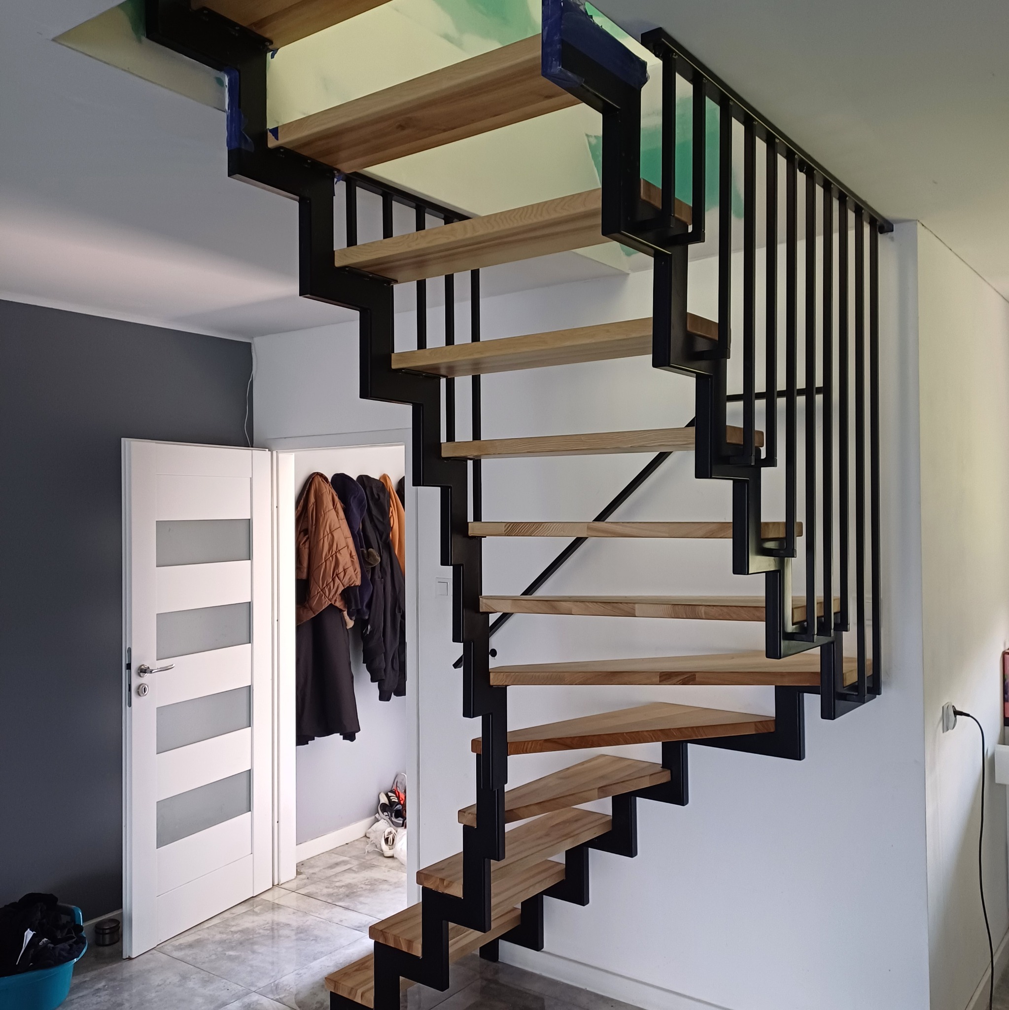 Nowoczesne schody metalowe z drewnianymi stopniami, czarna konstrukcja, minimalistyczny design, widok z dołu, jasne wnętrze, drzwi w tle.