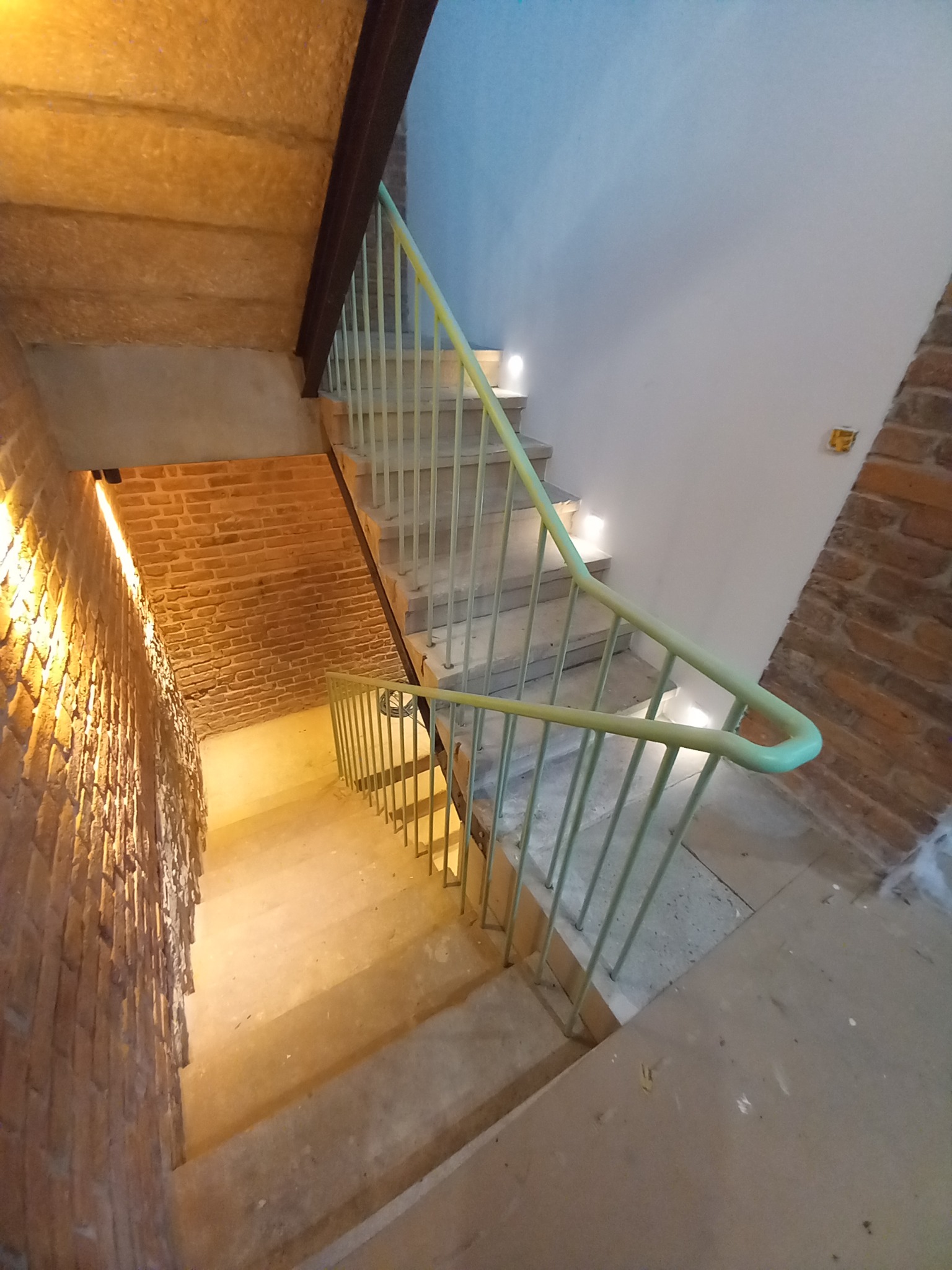 Betonowe schody z jasnozieloną poręczą w Świerzawie, oświetlone punktowymi lampami. Ceglana ściana z boku dodaje surowego charakteru wnętrzu.