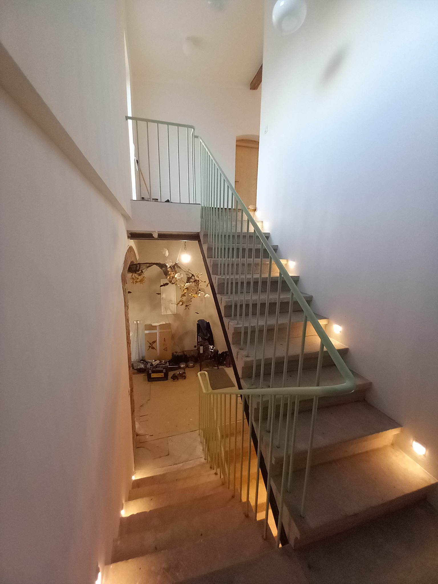 Schody betonowe z jasną, metalową balustradą i oświetleniem schodów. Widok z góry na dół, w tle widoczne narzędzia i materiały budowlane.