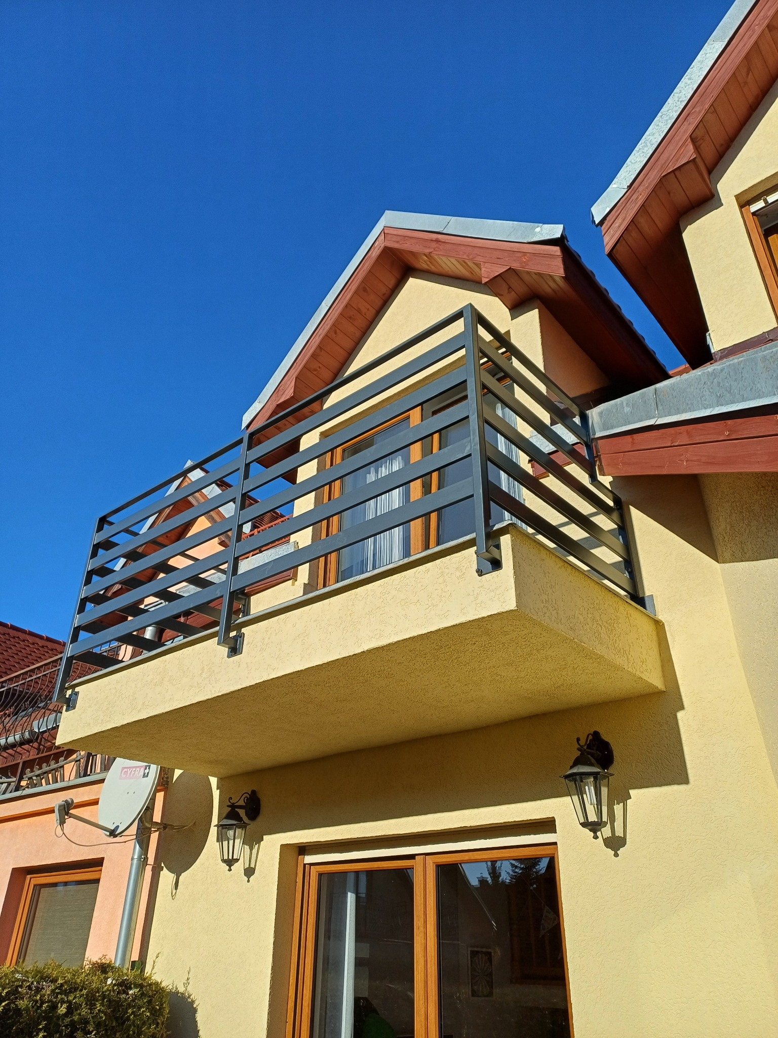 Balkon z nowoczesną, szarą balustradą z poziomych profili na tle błękitnego nieba i elewacji budynku w ciepłym odcieniu. Widoczne lampy zewnętrzne i fragment dachu.