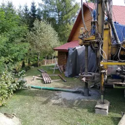 Żółty wiertniczy pojazd na trawie w pobliżu drewnianego domu z czerwoną dachówką podczas wiercenia otworu; widoczne rury wiertnicze, zielony wąż i błoto.