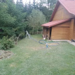 Wiercenie studni na trawniku obok drewnianego domku, widoczny sprzęt wiertniczy i sterta ziemi.