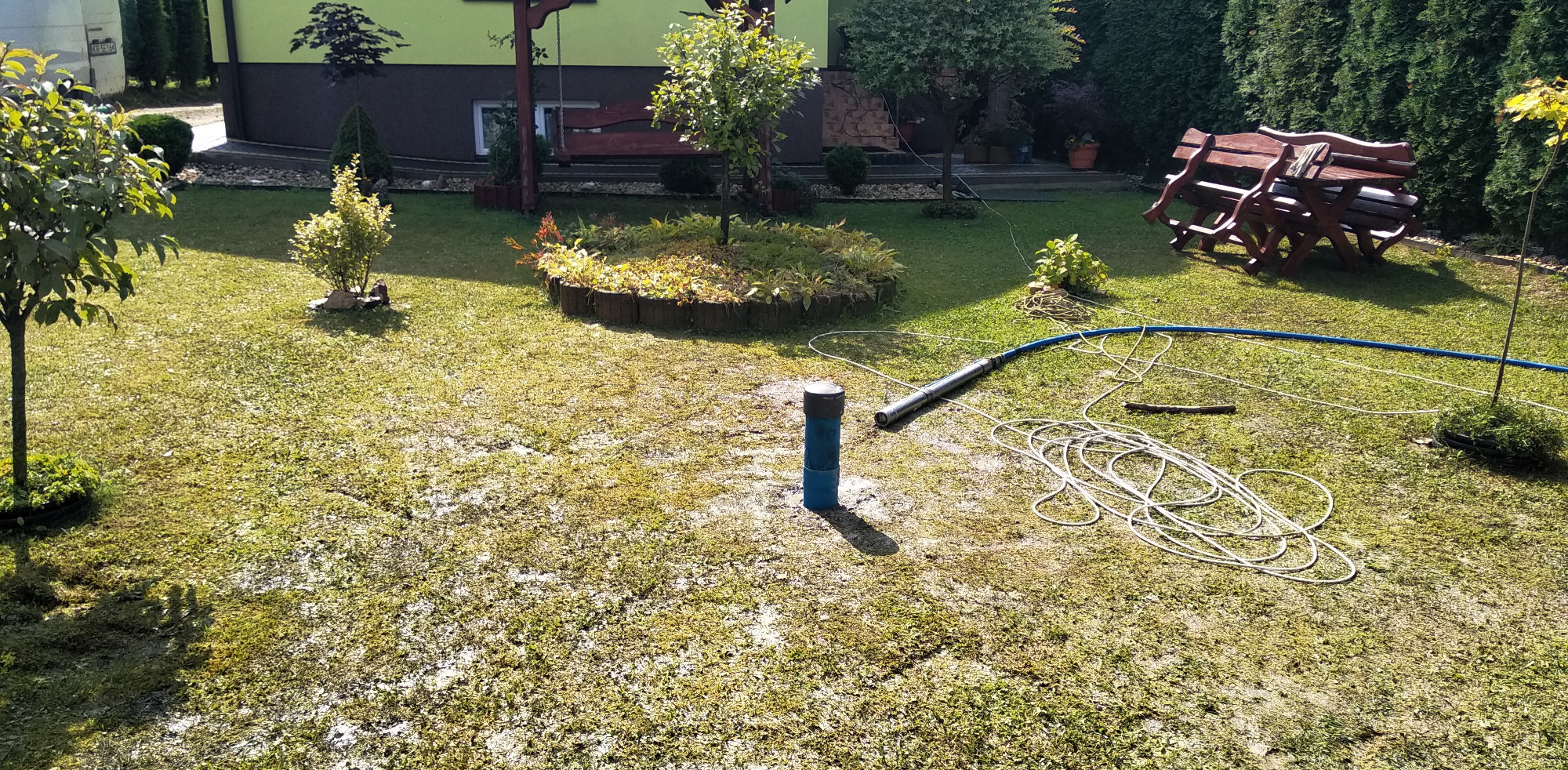 Niebieska rura wystająca z ziemi na trawniku, obok leży zwinięty biały kabel i niebieski wąż oraz pompa głębinowa, w tle widoczny ogród i dom.