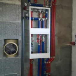 Rozdzielacz instalacji wodnej i ogrzewania podtynkowy w białej obudowie, z widocznymi rurami w kolorze czerwonym i niebieskim, zamontowany na surowej ścianie z betonu komórkowego. Dodatkowo...