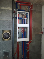 Rozdzielacz instalacji wodnej i ogrzewania podtynkowy w białej obudowie, z widocznymi rurami w kolorze czerwonym i niebieskim, zamontowany na surowej ścianie z betonu komórkowego. Dodatkowo...