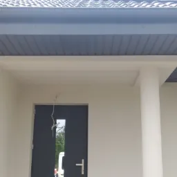 Fragment elewacji domu z szarym dachem krytym dachówką, widoczny fragment ocieplonej ściany, kolumna i czarne drzwi wejściowe z małym okienkiem, zwisający kabel przy drzwiach.