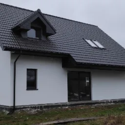 Nowoczesny dom jednorodzinny z białą elewacją, ciemną dachówką i oknami, widok z boku na tle pochmurnego nieba i jesiennego ogrodu.