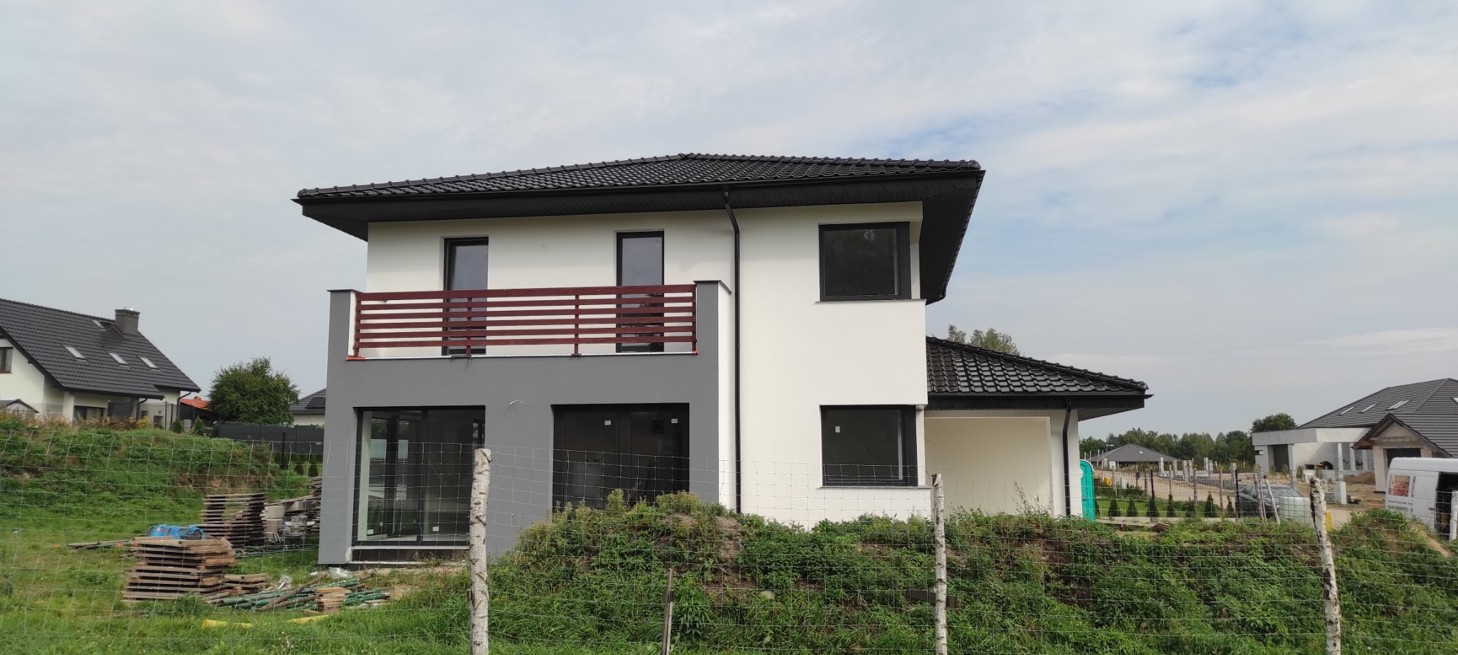 Dwukondygnacyjny dom z płaskim dachem i balkonem z drewnianą balustradą, elewacja częściowo w kolorze szarym i białym, widok z zewnątrz na tle innych domów i zieleni.