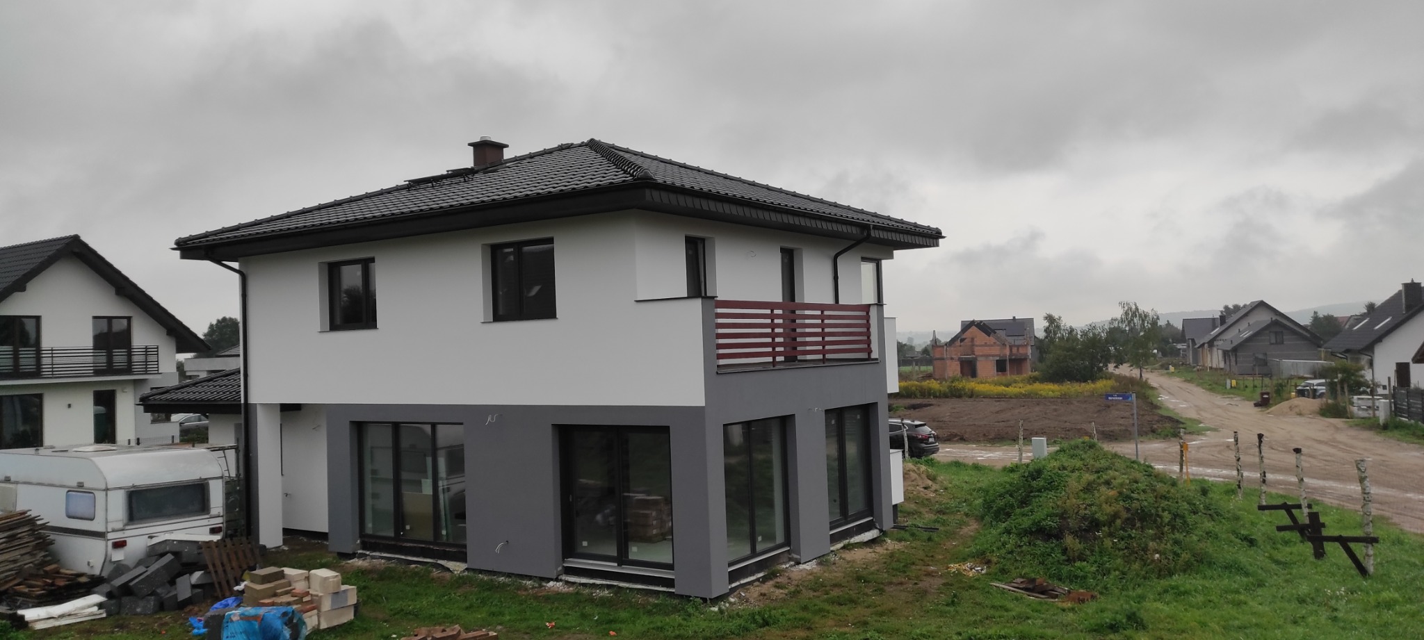 Nowoczesny dom dwukondygnacyjny z szarą dolną częścią elewacji i białym kolorem na górze, z balkonem z drewnianą balustradą, na tle pochmurnego nieba i innych domów w okolicy.