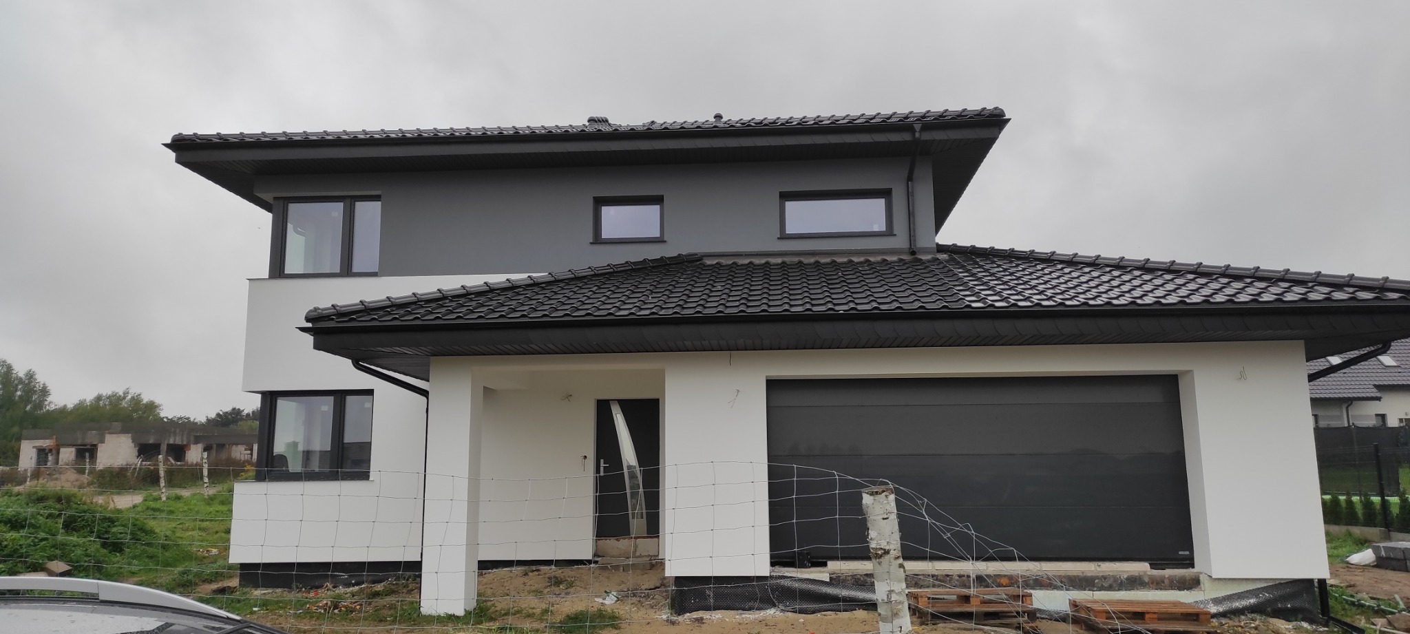 Nowoczesny dom jednorodzinny z szarą elewacją i czarnym dachem w Białogardzie, widoczny zza ogrodzenia z siatki, w tle niedokończone budynki.