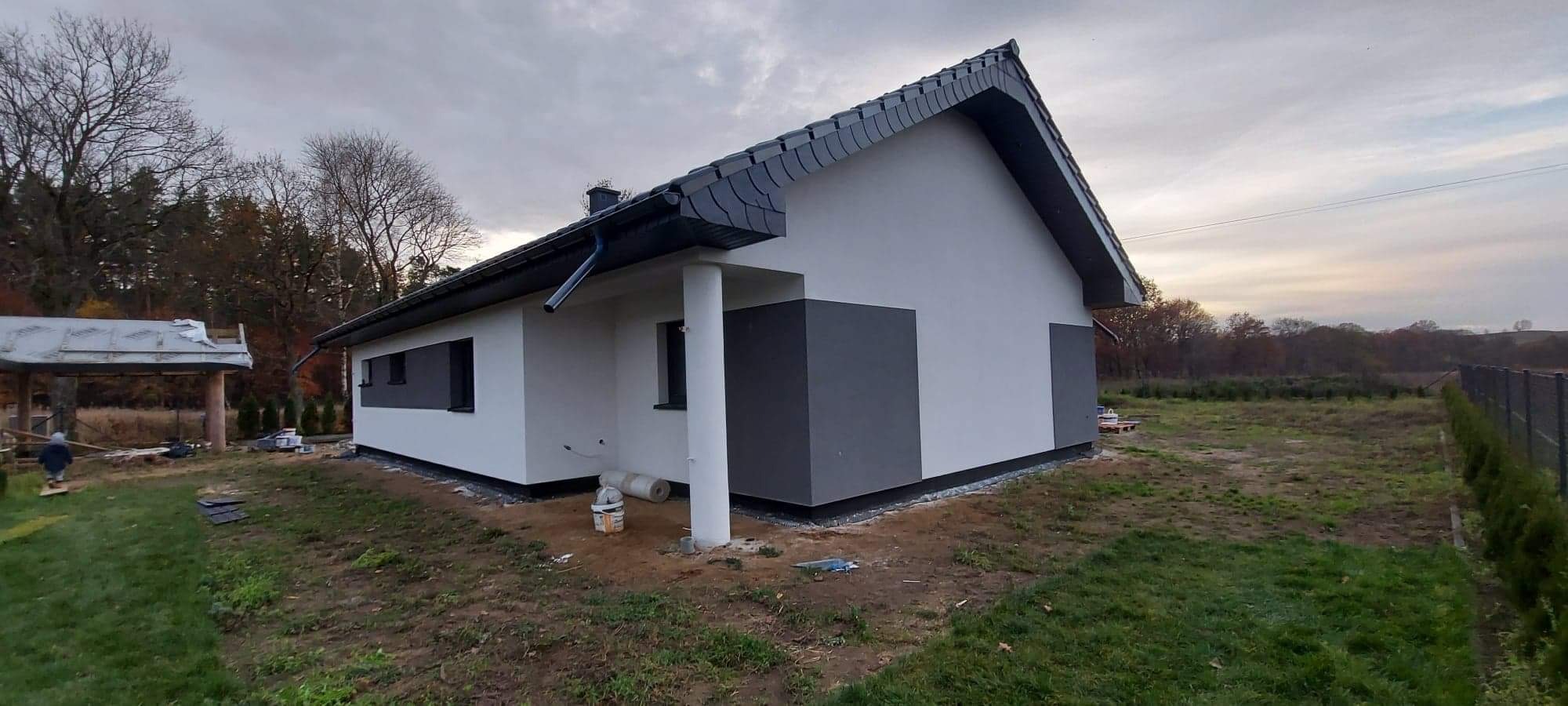 Nowoczesny dom jednorodzinny z elewacją w odcieniach szarości i bieli, widok z boku na częściowo zagospodarowaną działkę, z widocznym rusztowaniem w tle i materiałami budowlanymi przy ścianie.
