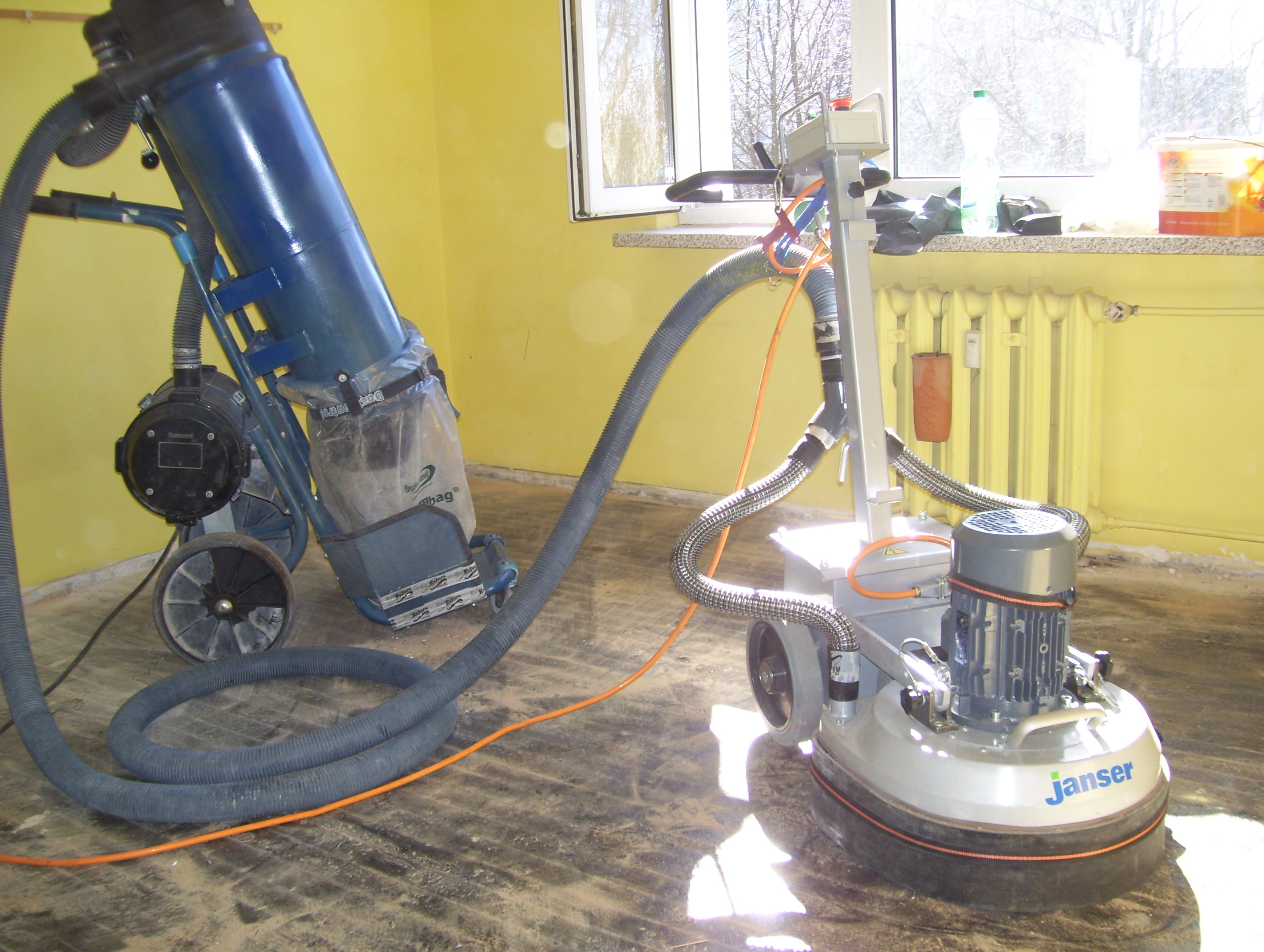 Parkiet szlifowany maszyną Janse, z podłączonym odkurzaczem przemysłowym, widoczne ślady po szlifowaniu, żółte ściany i okno w tle.