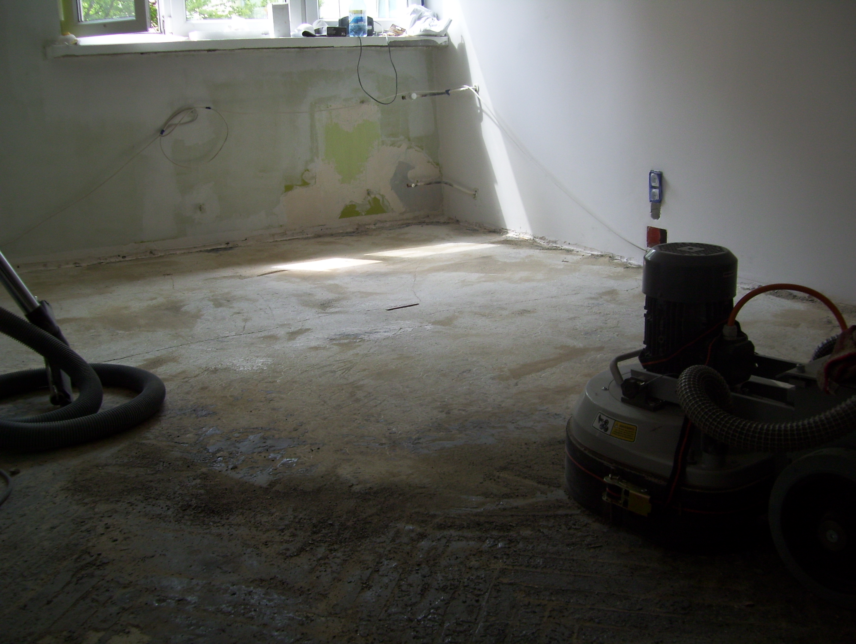 Przygotowanie podłoża pod nową podłogę: widok na pokój z resztkami starej wylewki, szlifierką do betonu i odkurzaczem przemysłowym.