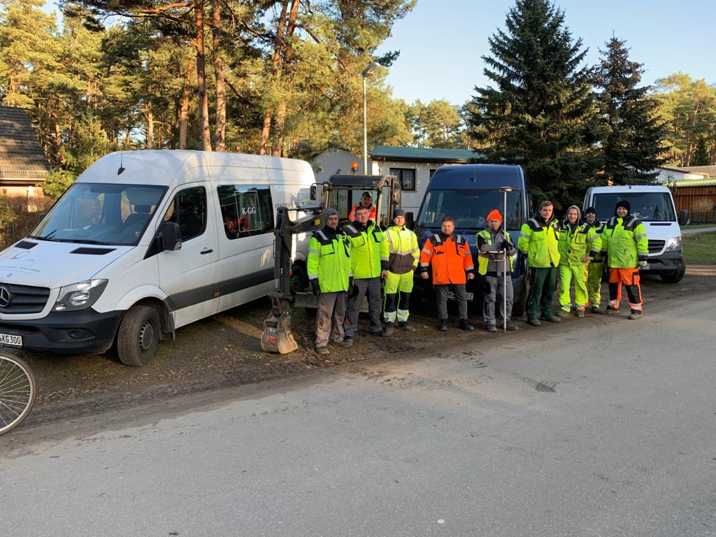 Grupa pracowników w odblaskowych ubraniach stoi przed trzema busami i małą koparką na ziemnej drodze, otoczona drzewami.