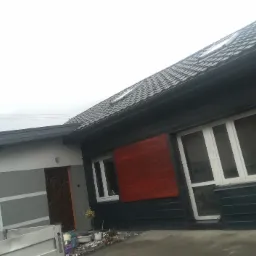 Dom jednorodzinny z ciemną dachówką i oknami w białych ramach, częściowo wykończony czerwoną drewnianą boazerią, widok z poziomu gruntu