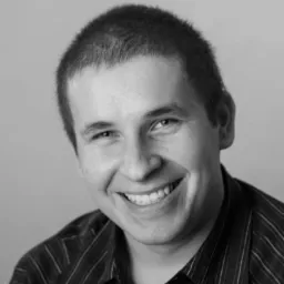 Czarno-biały portret uśmiechniętego mężczyzny w koszuli w paski, z widocznymi zmarszczkami mimicznymi wokół oczu i charakterystycznym układem zębów.