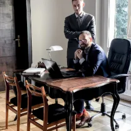 Dwóch prawników w garniturach w biurze: jeden siedzi za biurkiem z laptopem, drugi stoi za nim z założonymi rękami, widoczne pomarańczowe skarpetki i brązowe buty siedzącego adwokata.