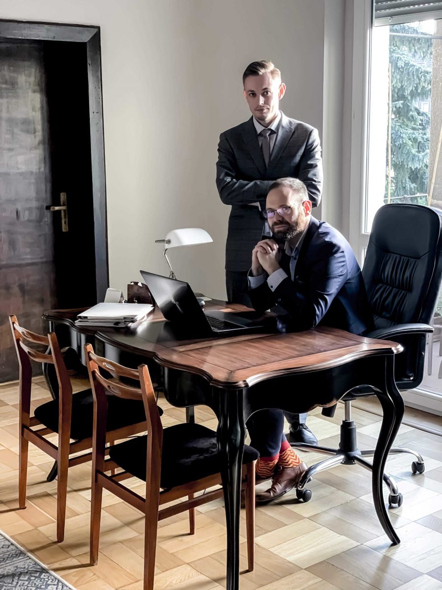 Dwóch prawników w garniturach w biurze: jeden siedzi za biurkiem z laptopem, drugi stoi za nim z założonymi rękami, widoczne pomarańczowe skarpetki i brązowe buty siedzącego adwokata.