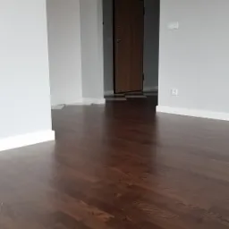 Wnętrze mieszkania po remoncie z ciemną, drewnianą podłogą ułożoną w jodełkę, szare ściany i widok na przedpokój z brązowymi drzwiami wejściowymi.