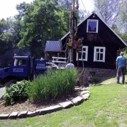 Wiercenie studni przy drewnianym domu na wzgórzu, widoczny specjalistyczny samochód z napisem 'Studnie Wiercone' i ekipa w trakcie pracy.